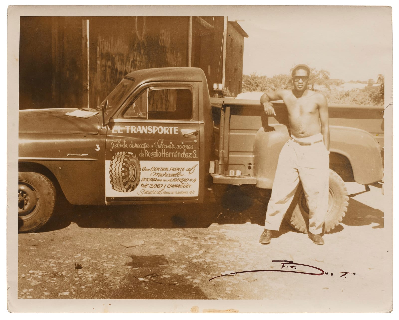 Sepia photo of a topless man resting his arm on the back of a truck that has hand-painted lettering on the door.