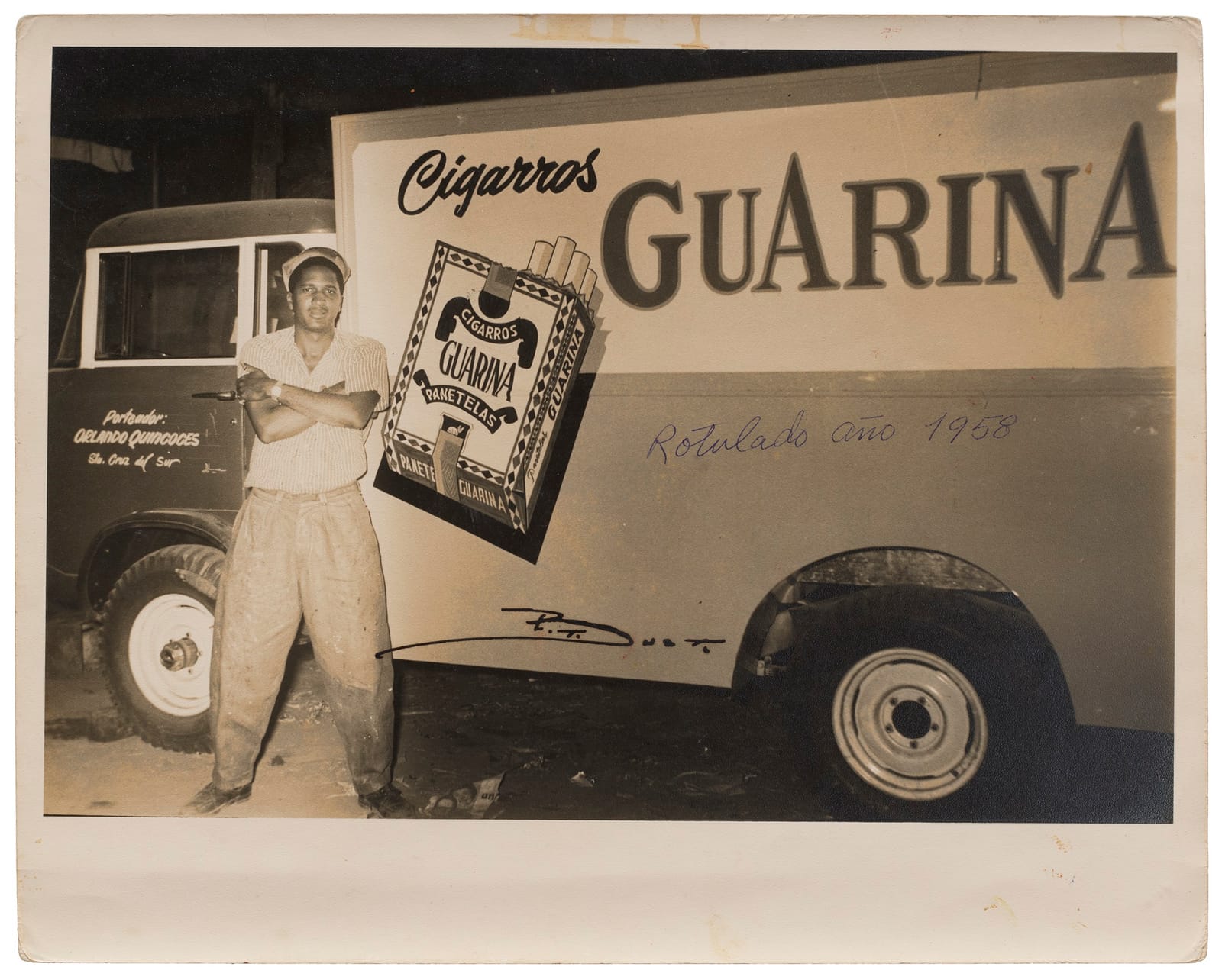 Smartly dressed man with arms folded in front of a lorry that has a picture of a packet of cigars and lettering for the Guarina brand.