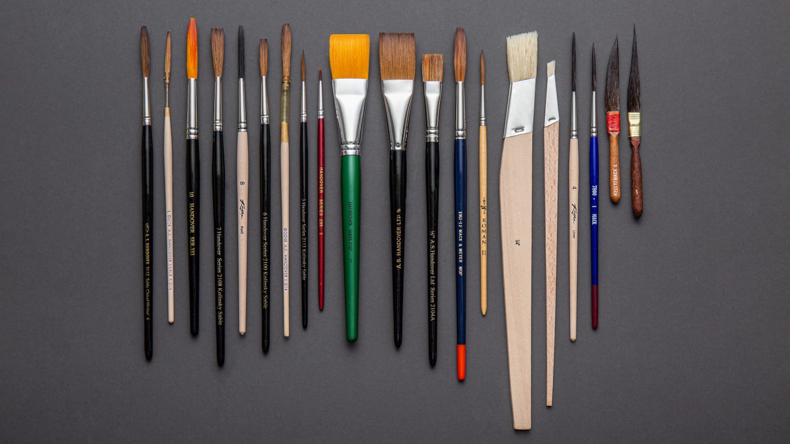 20 different types of brushes lined up on a surface and photographed from above so that their handles and hairs are visible.