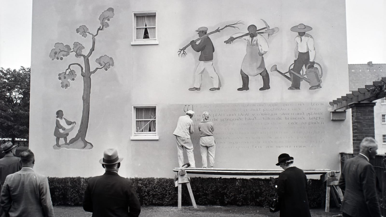 Black and white photo of two sign painters on a tressle working on small lettering that makes up a large paragraph of text on a wall. Above that portion of the wall are three painted agricultural workers, and further to the left is a woman standing under a tree.