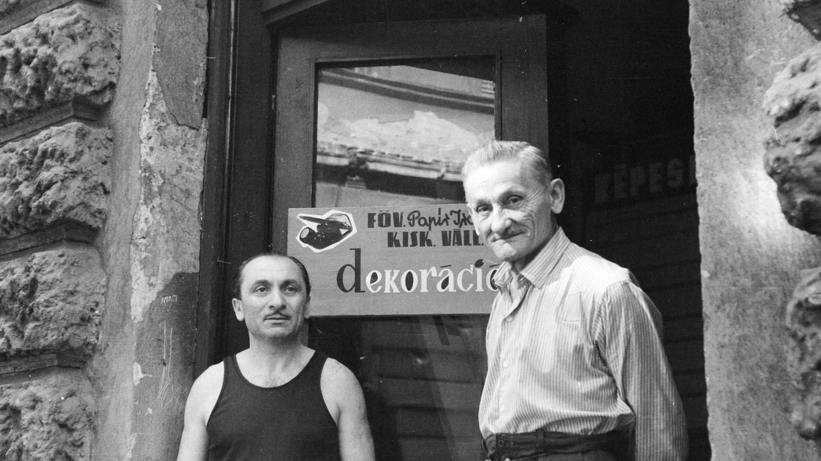 Black and white portait of two men, one older than the other, standing in the doorway of a shop. There is a sign hanging on the door which isn't fully visible, but with has the word 'dekoracion' on it, suggesting that they are decorators/sign painters.