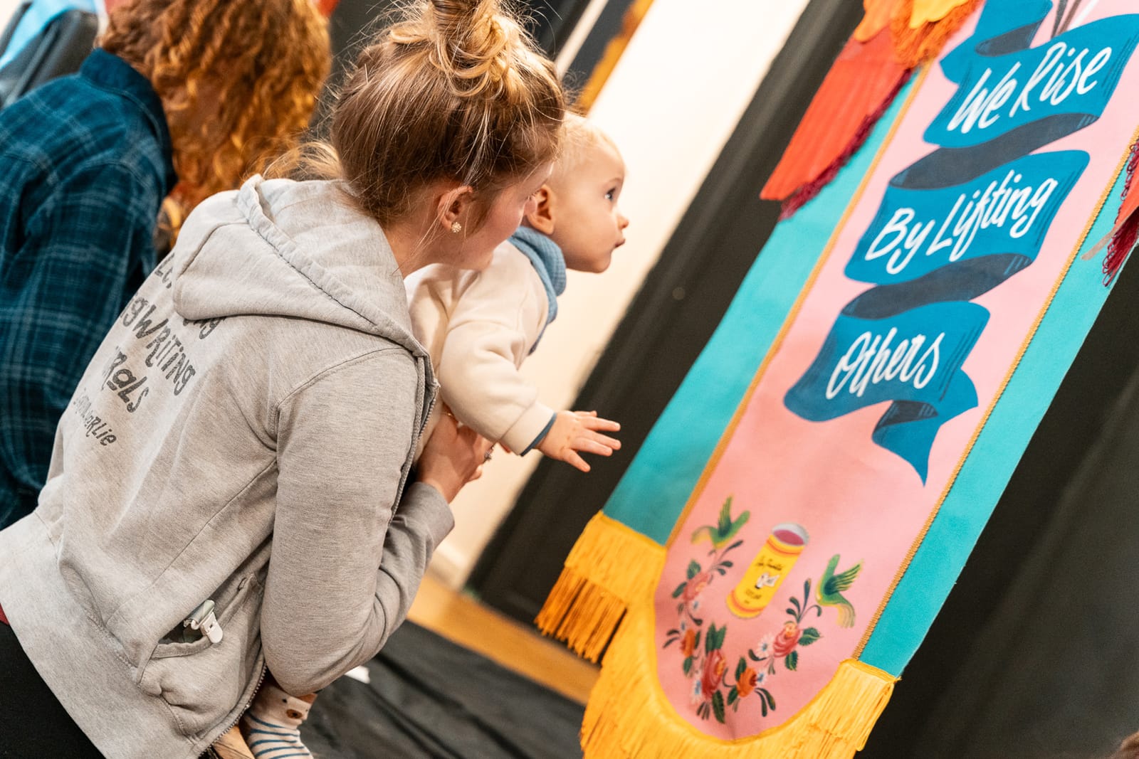 A woman holding a baby that's looking at painted lettering that says 'we rise by lifting others'.