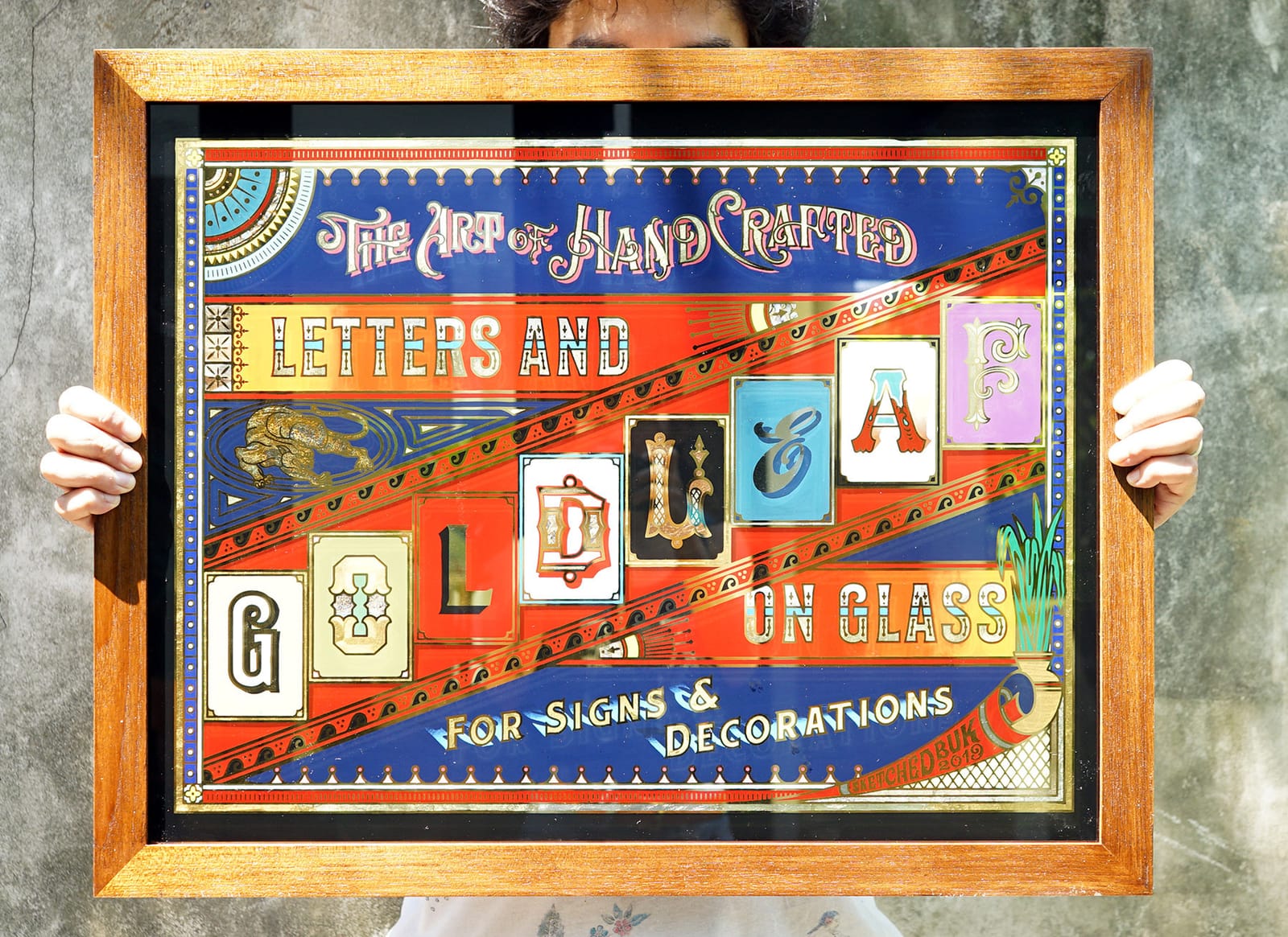 Man holding a framed glass piece that uses gilding, sign painting, and other decorative elements to demonstrate their appearance with the wording, "The Art of Hand-Crafted Letters and Gold Leaf on Glass For Signs & Decorations".