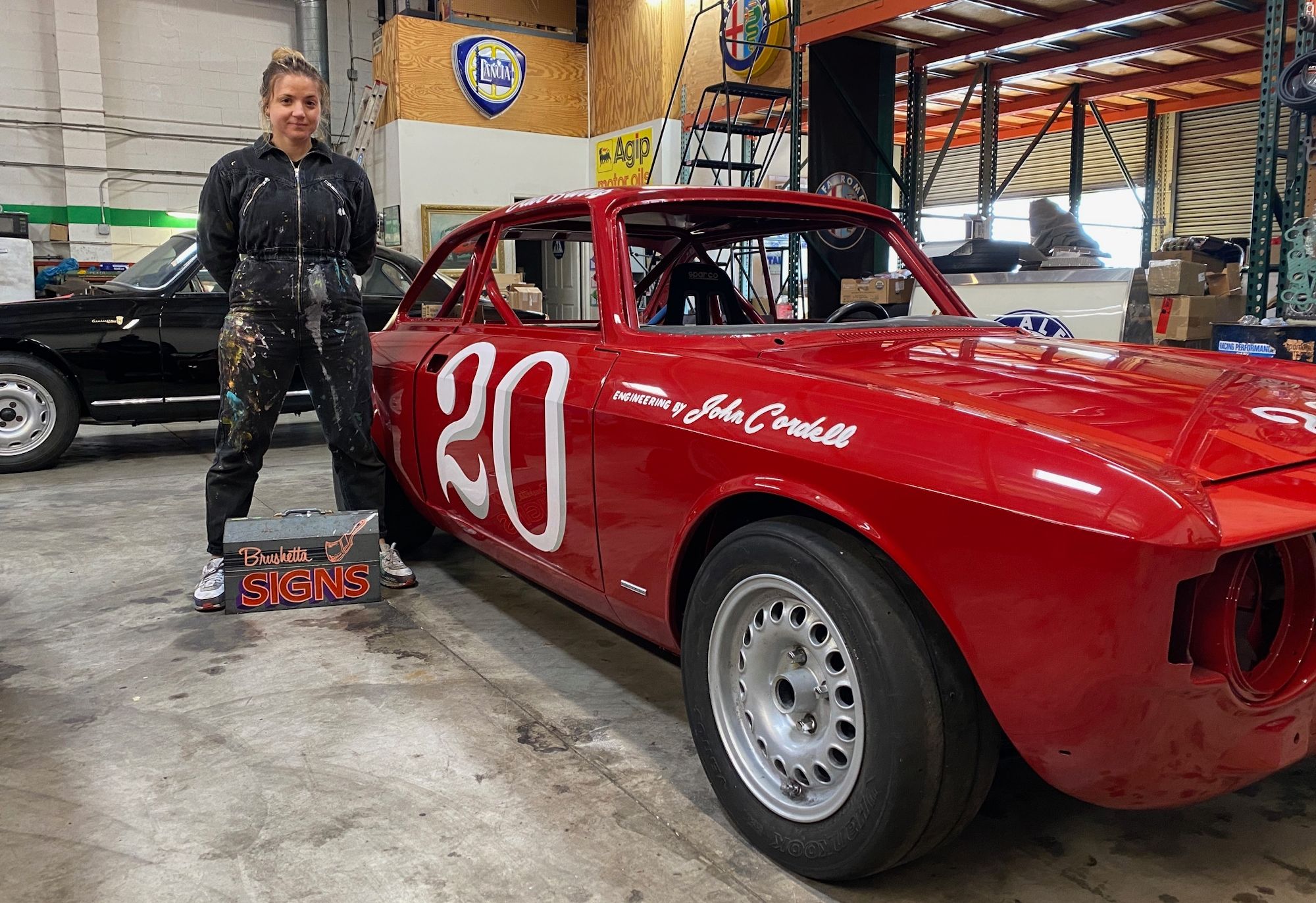 Woman in overalls and sign painting kit standing next to a bright red racing car painted with the number 20 on its side.