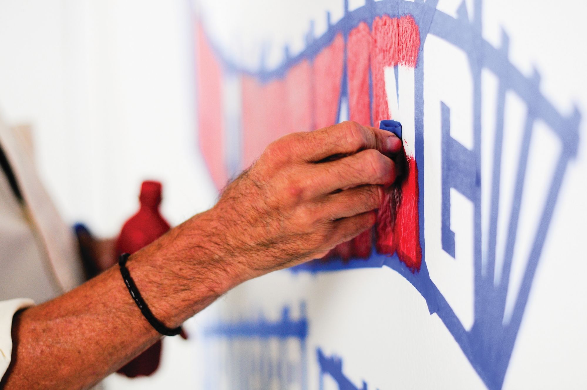 Close-up of a hand applying paint to a wall with letters outlined in masking tape.