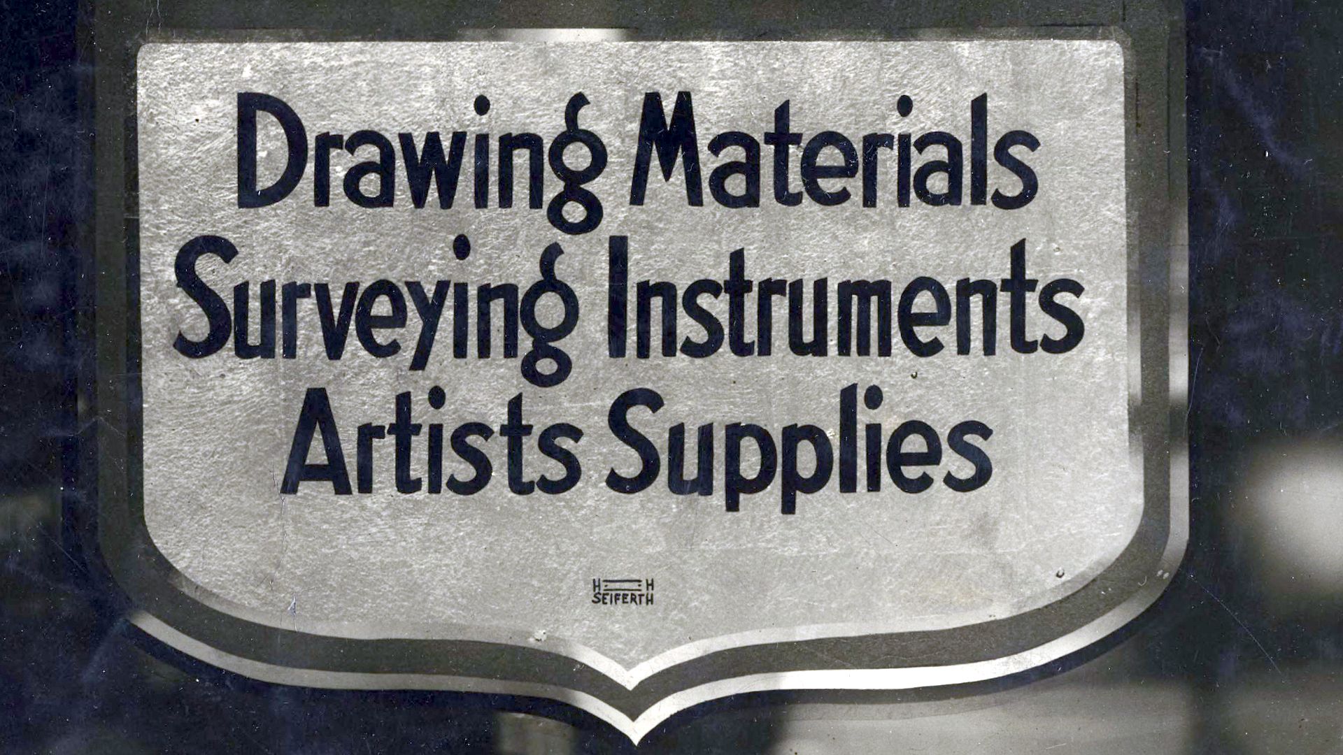 Plaque-shaped sign with hand-painted words, "Drawing materials, surveying instruments, artists supplies".
