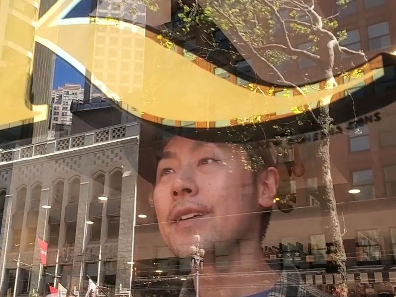 Man looking through a window with photo taken from the outside. Above is part of a gilded letter K.