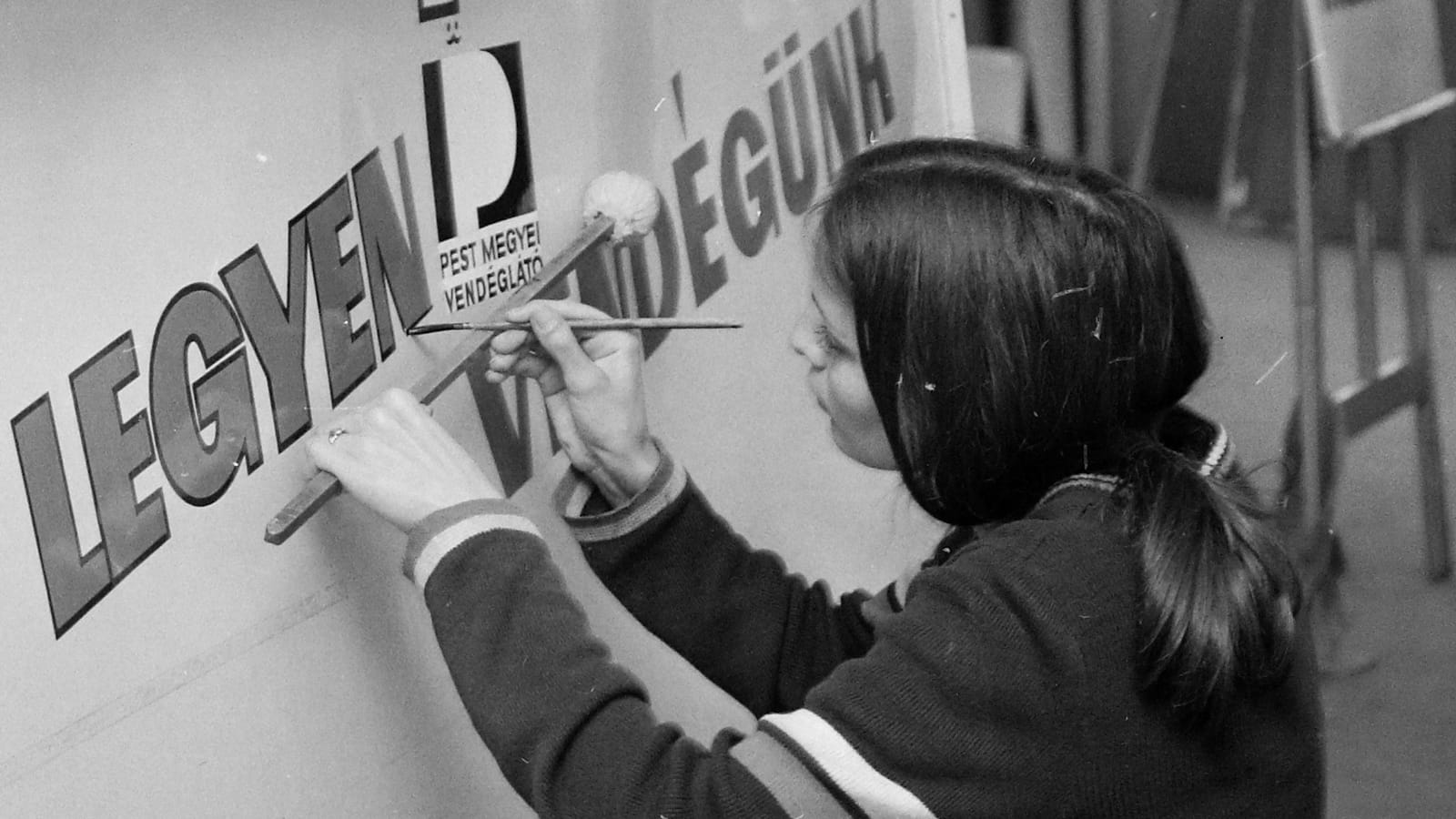 Over-the-shoulder view of a woman holding a mahl stick and painting lettering on a sign board.