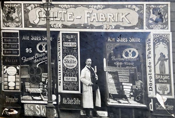 Archival photo of a sign painter standing at the entrance to his shop with is adorned with numerous signs.