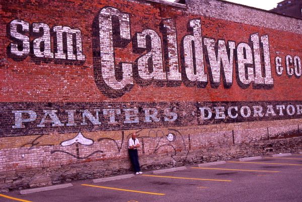 Elderly man standing in front of an enormous fading painted sign advertising Sam Caldwell & Co., Painters & Decorators..