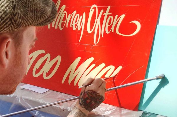 Man with a brush in his hand, resting it on a mahl stick, and painting cream lettering onto a red panel.