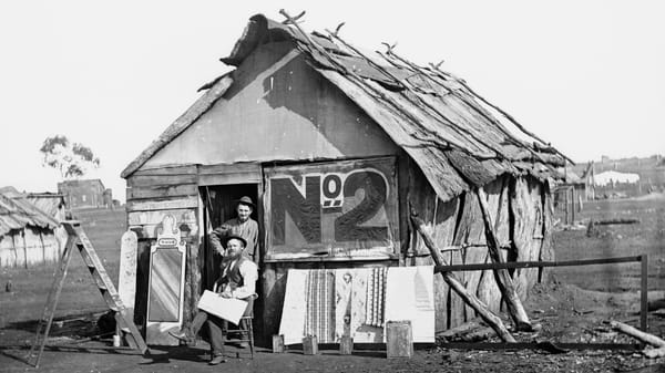 Two men posing outside a ramshakle timber shack with a large No.2 painted on the side, and bits of sign painting work around.