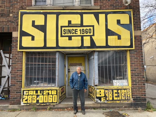 Elderly man standing in front of a shopfront which is emblazoned with a huge fascia sign that reads "SIGNS".
