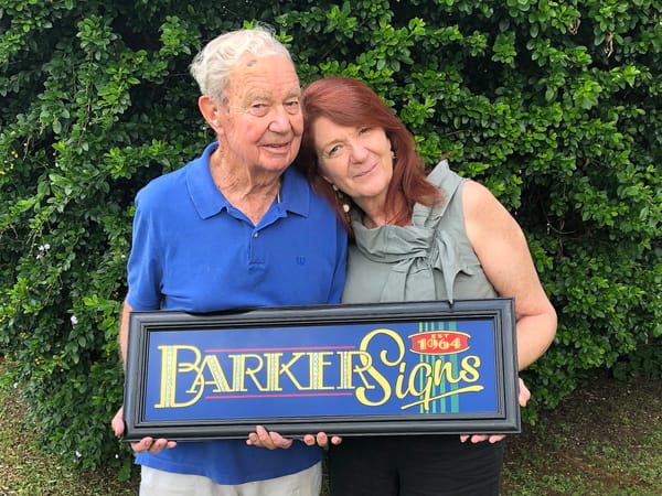 Man and woman facing the camera, holding a framed sign that reads "Barker Signs, Est. 1964".