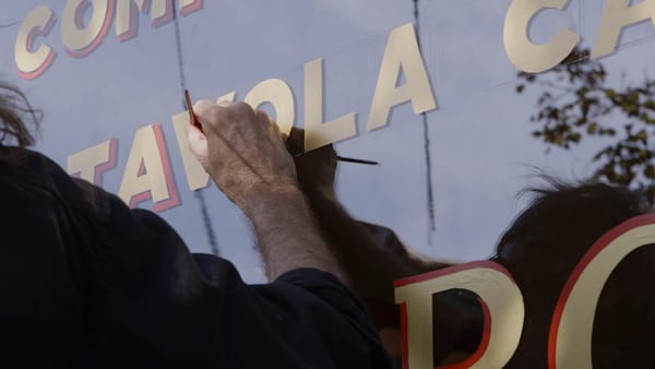 View of a hand holding a brush painting a red shadow on golden block letters on a window.