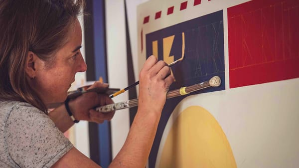 View from the side of a woman leaning on a rest (mahl) stick and painting some lettering in yellow on a blue panel.