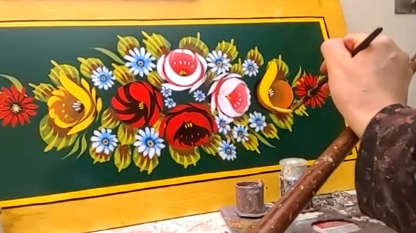Close cropped photo of a hand resting on a mahl stick painting a bouquet of roses on a rectangular panel.