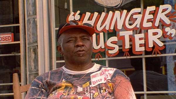 Head and shoulders view of a man in a baseball cap in front of a window sign that reads "Hunger Buster".