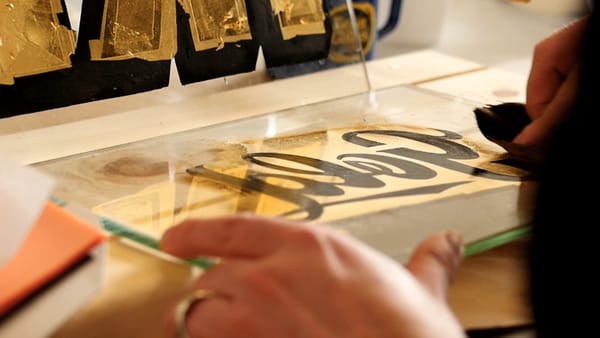 View of a hand holding a glass panel with shiny gold leaf and black lettering on top that, in reverse, reads 'Gold'.