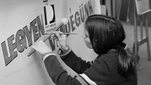 Over-the-shoulder view of a woman holding a mahl stick and painting lettering on a sign board.