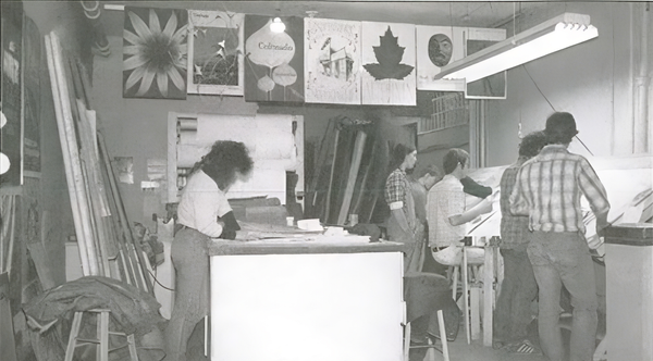 Black and white photo of a small group of people in a studio setting, engrossed in drawing and conversation.