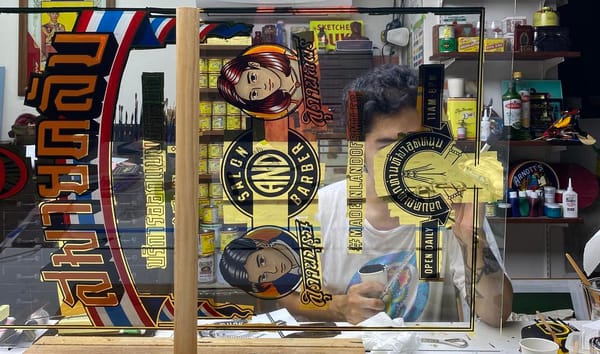 Man working behind a sheet of glass adorned with gilded and painted lettering and pictorials.