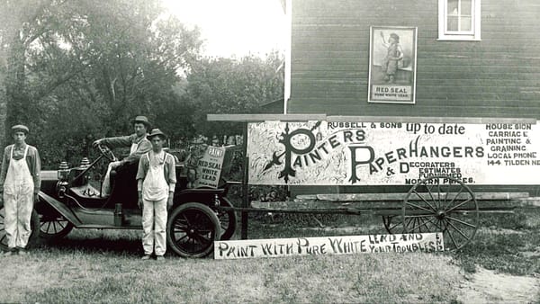 Men in overalls posing on/in front of an old car that's hitched to a large trailer adorned with a sign advertising their business.