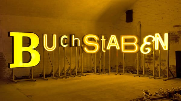 Diverse letters with yellow illumination of different forms arranged on supported stands to spell 'Buchstaben'.