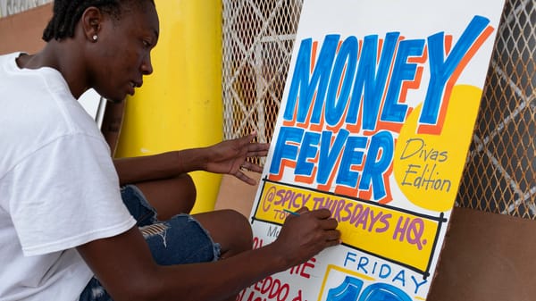 Man sat on a stool painting a sign advertising "Money Fever, Divas Edition, at Spicy Thursdays HQ."