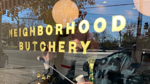 Sign painter working on the inside a window with gilded letters that read "Neighbourhood Butchery".