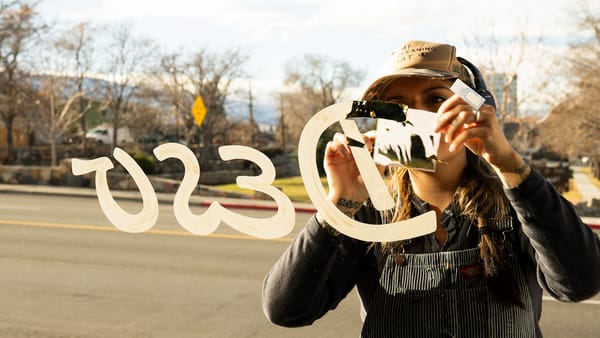 View through a window as a sign painter applies gold leaf to the lettering painted on the glass. The text is in reverse and reads "Desu".