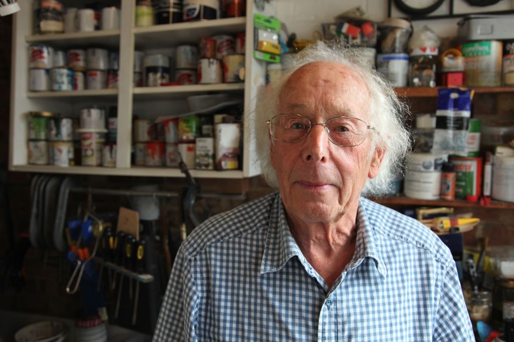 Portrait of elderly man with shelves of paints and solvents behind him.
