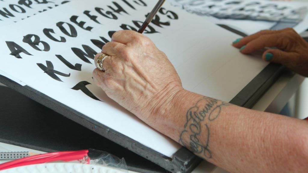 Close-up of left hand painting lettering in black on white paper.